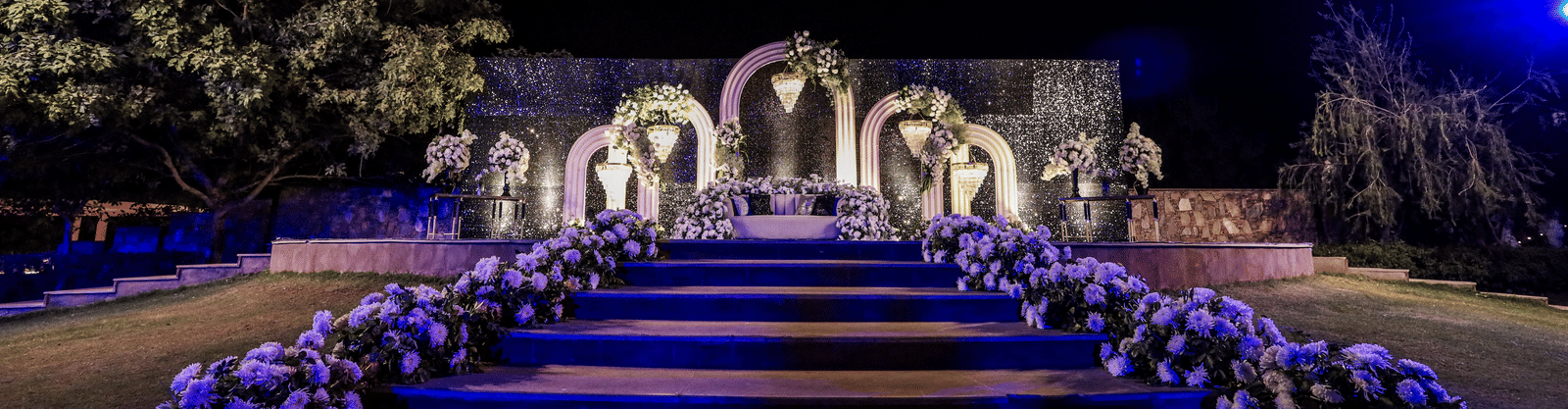 A wedding stage at Ananta Spa & Resort, Jaipur, is viewed across blue-lit steps adorned with purple and white flowers at night.