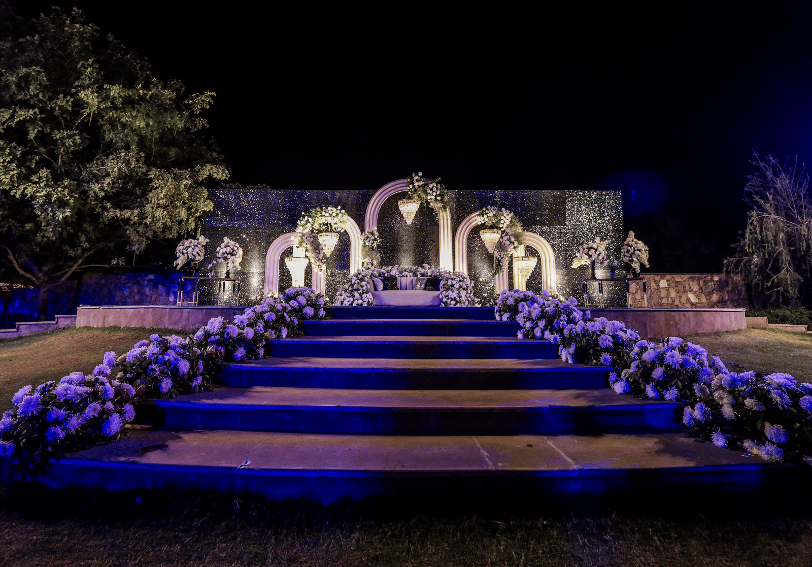 A wedding stage at Ananta Spa & Resort, Jaipur, is viewed across blue-lit steps adorned with purple and white flowers at night.