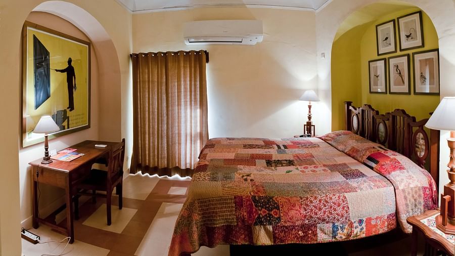 Bedroom with a bed featuring a patchwork quilt, bedside tables with lamps, a desk, artwork, curtains, and AC at Neemrana's Tijara Fort Palace.