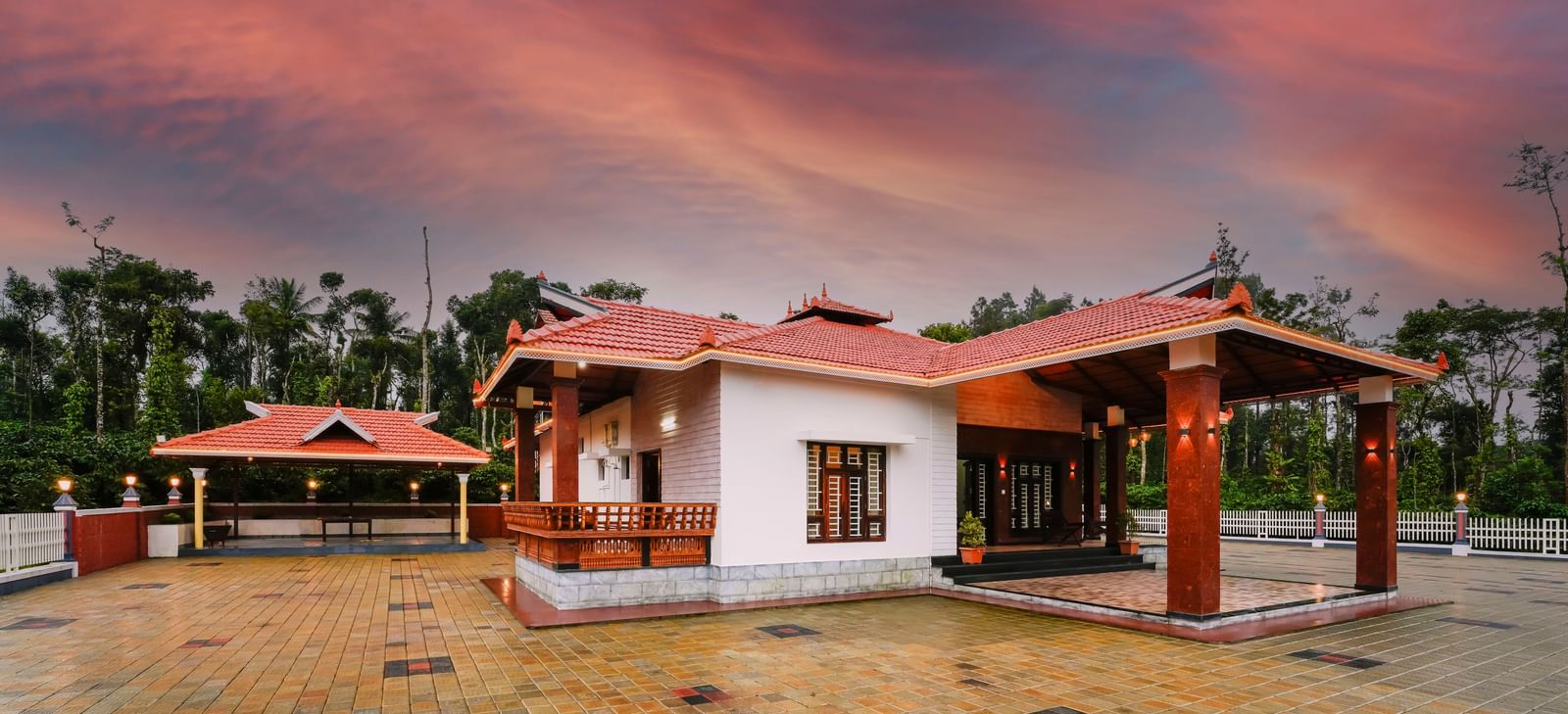A beautiful low-angle view of a cottage at Bhuthanakadu Retreat, featuring a red-tiled roof and a patio with rustic seating, all set against a stunning orange sunset.