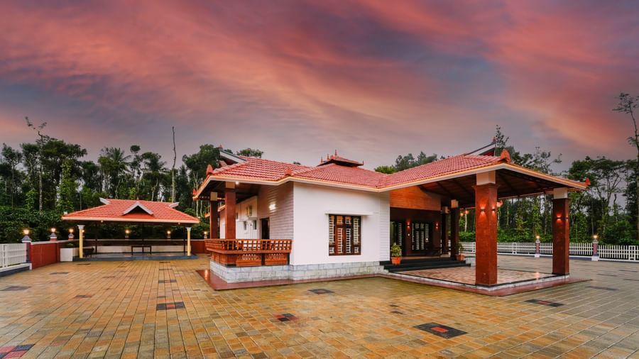 A beautiful low-angle view of a cottage at Bhuthanakadu Retreat, featuring a red-tiled roof and a patio with rustic seating, all set against a stunning orange sunset.