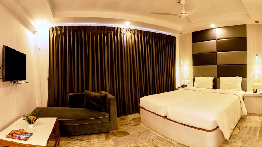 A comfortable and modern hotel room at Hotel Mamallaa Heritage, featuring a large bed with white linens, a television, and dark curtains.