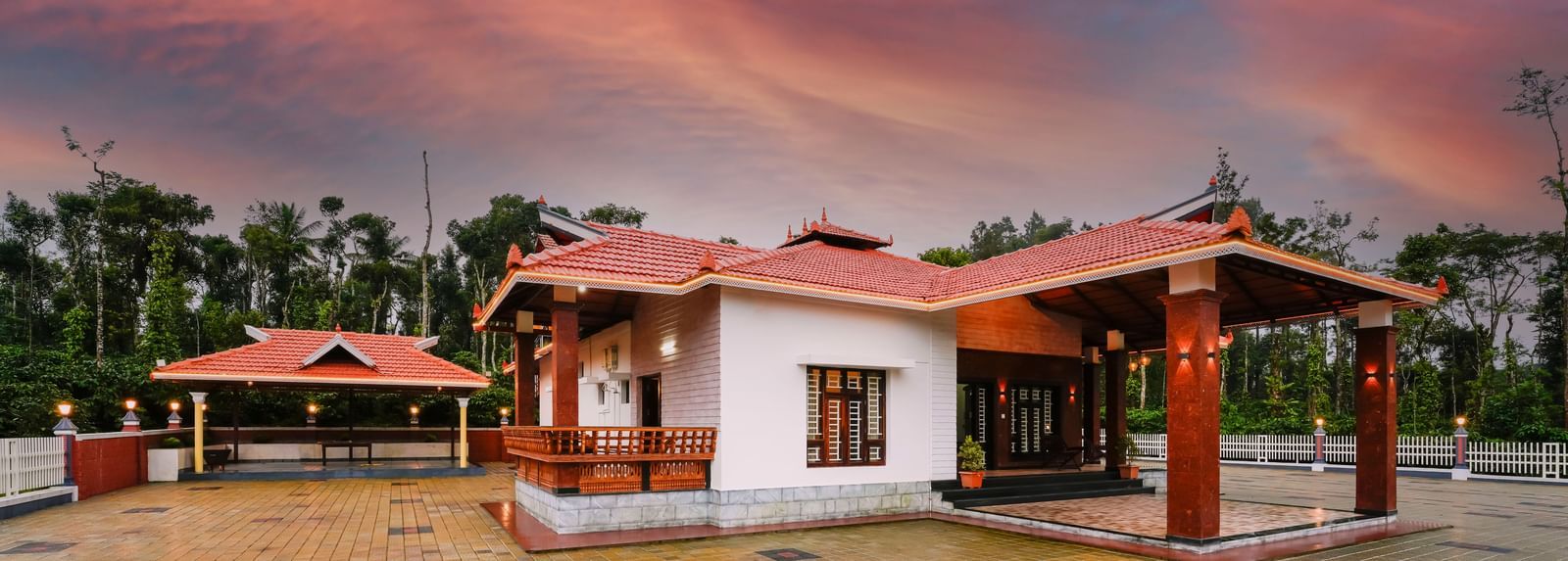 The traditional facade of Bhuthanakadu Retreat under a dramatic pink and orange sunset sky, showcasing the main building with its classic red-tiled roof.