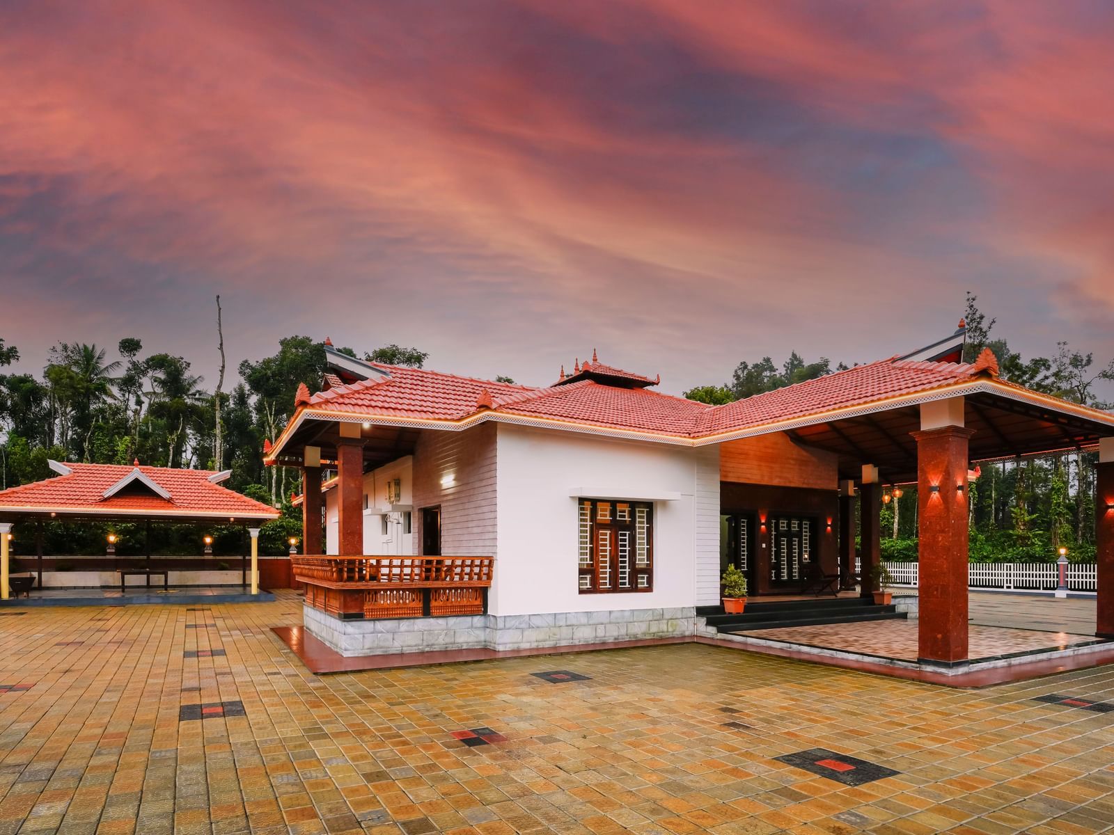 A beautiful low-angle view of a cottage at Bhuthanakadu Retreat, featuring a red-tiled roof and a patio with rustic seating, all set against a stunning orange sunset.
