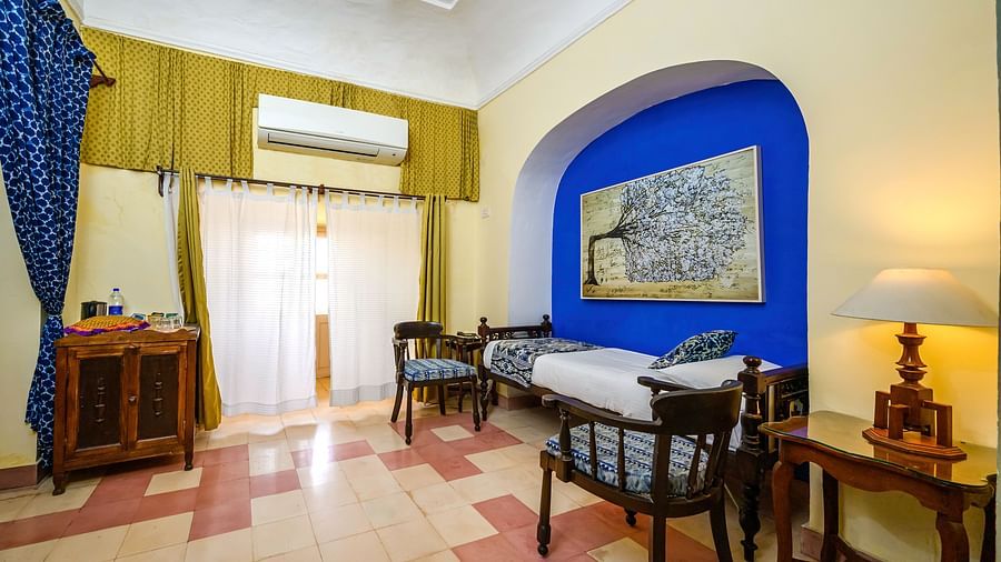 Room with bed, patterned pillows, furniture including chairs, tables, and a cabinet, curtains, wall-mounted air conditioner, artwork in arched alcove, and patterned floor tiles - Tijara Fort-Palace - 19th Century, Alwar.