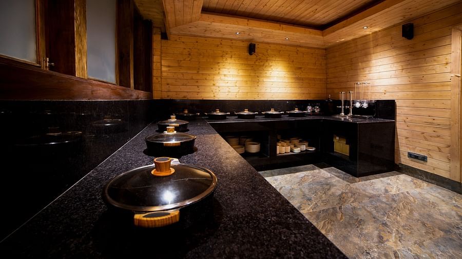 Modern buffet counter in a warmly lit wooden dining area, featuring a row of sleek black granite counters with covered serving dishes, matching black cabinetry, and natural stone flooring - Yashshree Lachen.
