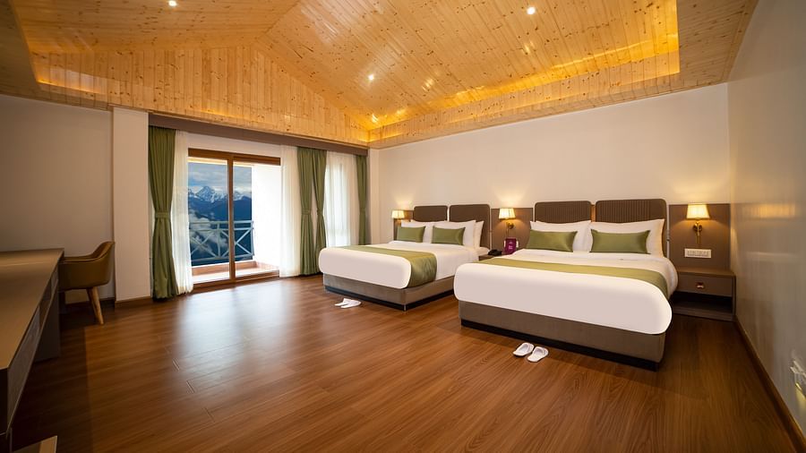Spacious hotel room with two large beds, wooden flooring, a desk and chair, warm ceiling lighting, and a balcony with a scenic view - Yashshree Pelling Chekhim Retreat.