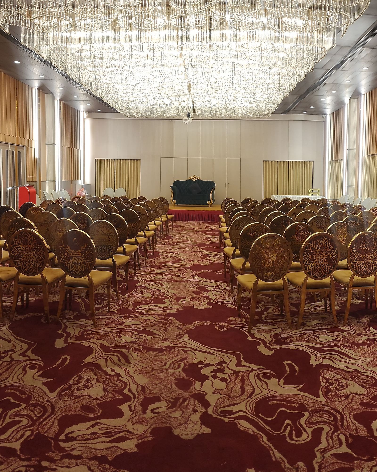 A spacious conference room with ornate chairs arranged in rows on a patterned red carpet, and a modern chandelier on the ceiling.