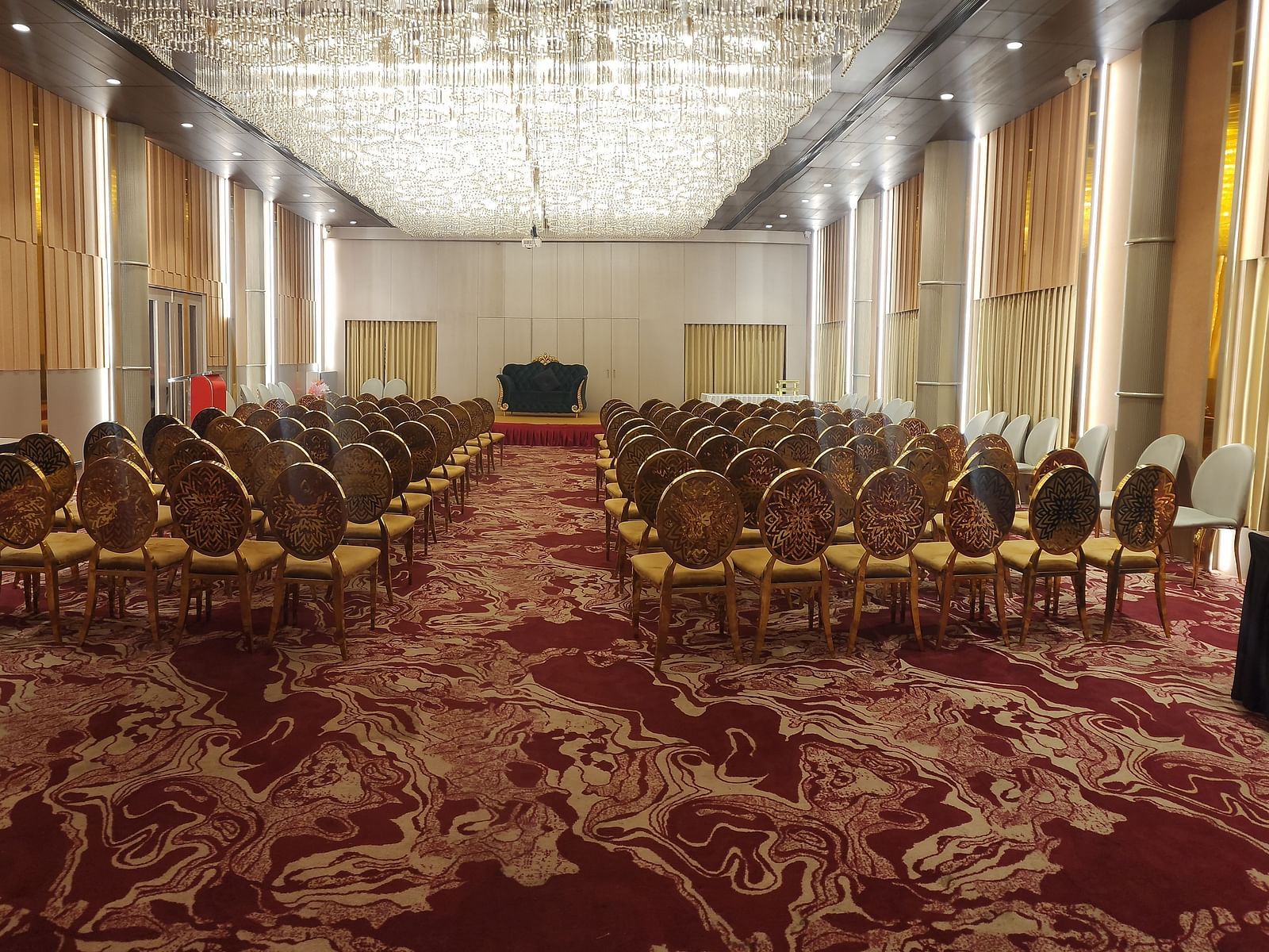 A spacious conference room with ornate chairs arranged in rows on a patterned red carpet, and a modern chandelier on the ceiling.