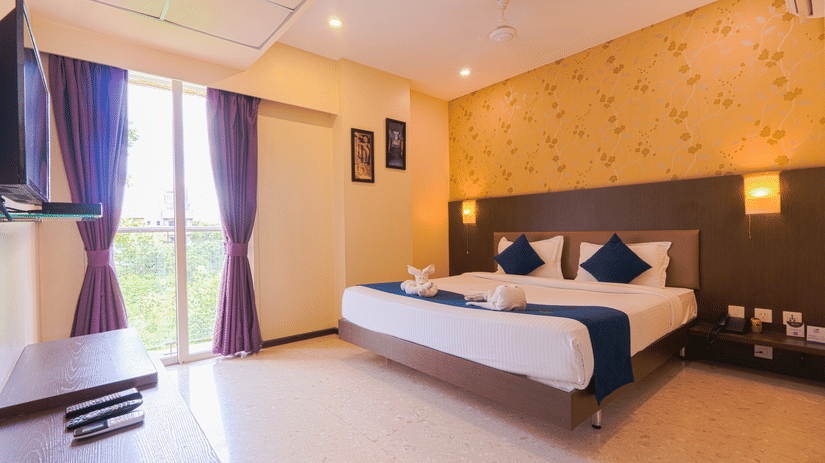 Guest room at Magnus Star Residency, Koregaon Park Annexe, featuring a large neatly made bed with decorative folded towels, pillows, soft lighting, wall art, a balcony with curtains, a mounted television, and a side table with remotes.