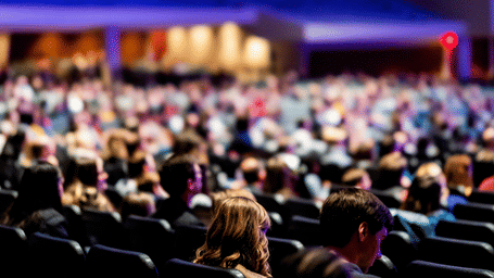 A large audience seated in a dimly lit auditorium during a conference or event, with stage lighting visible in the background