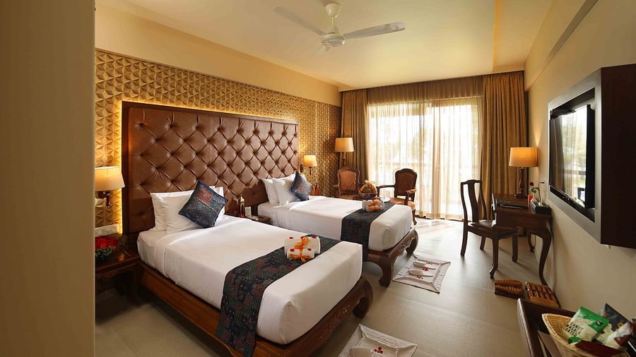 Lake View Deluxe Twin Room at Uday Backwater Resort featuring twin beds positioned next to each other in front of a wall-mounted TV, with a work desk nearby and a comfortable seating area by the window behind it