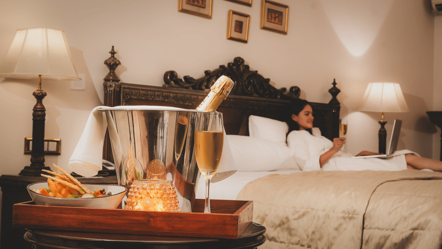 A bed with pillows and bedside lamps with a champagne bottle placed in an ice bucket on a tray in the foreground at The Suryaa, New Delhi.