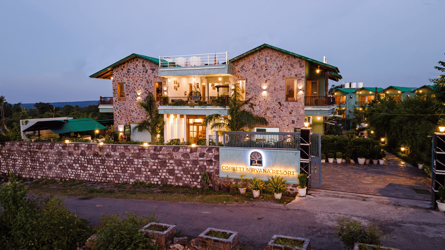 Well lit facade of Corbett Nirvana Resort, Uttarakhand featuring two storey cottage style building with balconies and terrace.