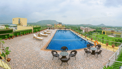 Rooftop swimming pool with blue water, patio furniture, and sun loungers overlooking a green landscape and hills under a cloudy sky at Golden Tulip Kukas, Jaipur.