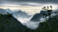 A view of serene mountains covered with fog and cloudy sky in the background.