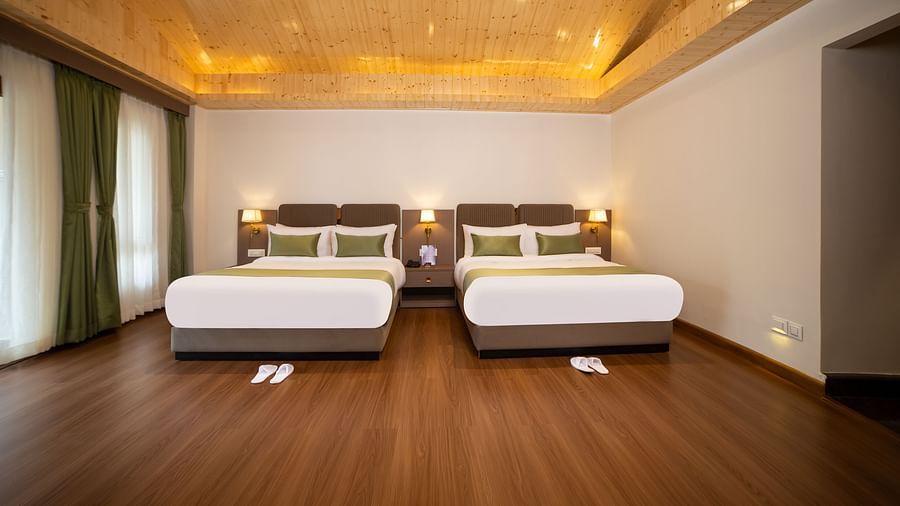 Spacious hotel room with two large beds featuring green and white bedding, wooden flooring, warm ceiling lighting, and large windows with curtains - Yashshree Pelling Chekhim Retreat.