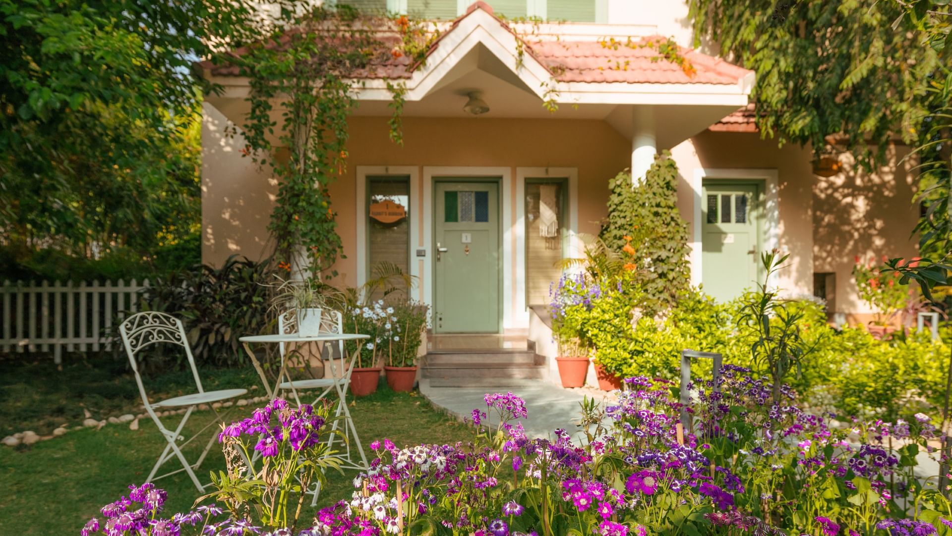 Charming villa entrance with pastel green doors, blooming purple flowers, potted plants, and outdoor seating at Karma Chalets.
