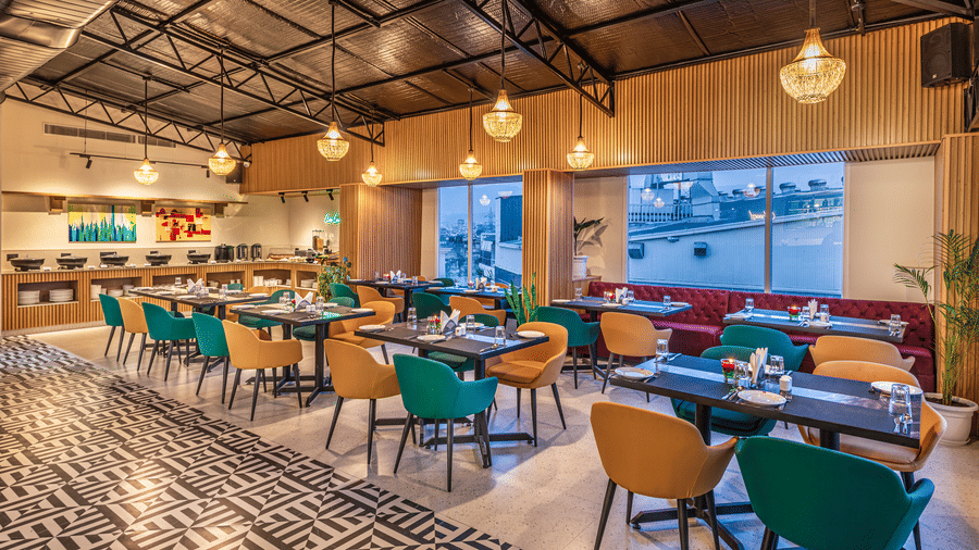 A modern restaurant with green and yellow chairs, dark tables, window view, and chandeliers and a buffet serving area at the background at Lords Eco Inn Dehradun.