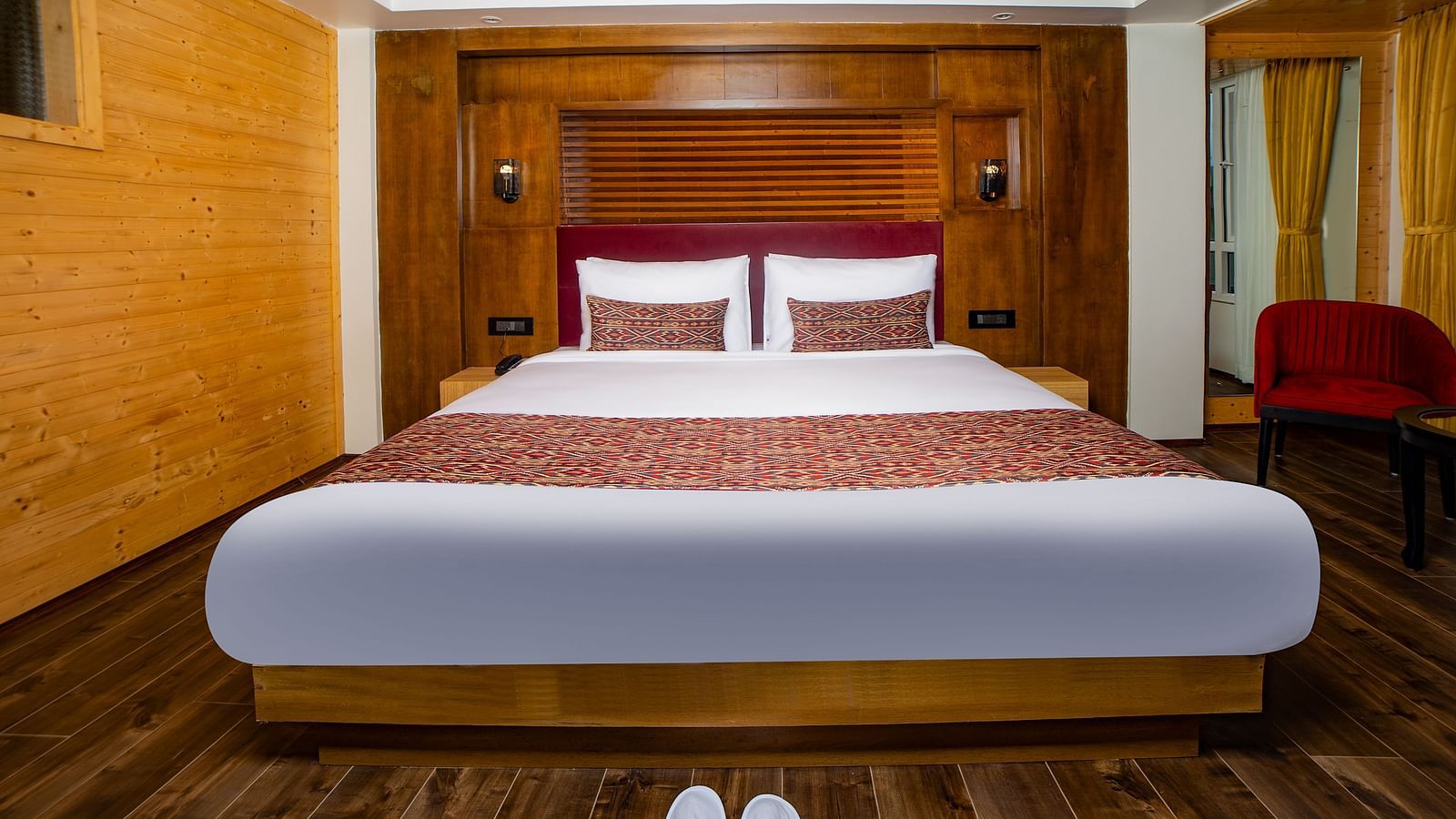 Cosy hotel bedroom with a large double bed featuring white linens and decorative red accents, set against a wooden-paneled wall, with wooden flooring, bedside lamps, and a red chair in the corner - Yashshree Lachen.
