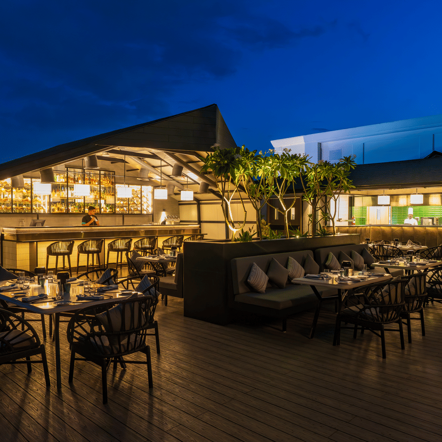 Evening view of The Residency Towers Puducherry's outdoor dining, tables set amidst lush greenery and warm lighting.