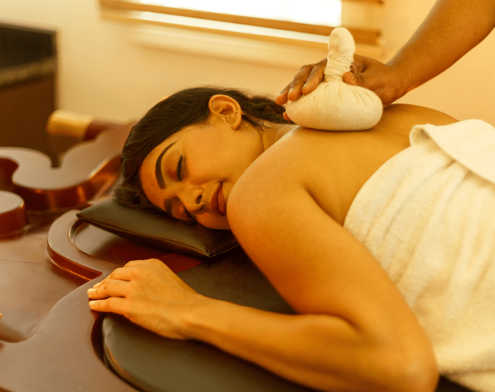 A person receiving a shoulder massage with a poultice while lying on a massage table.