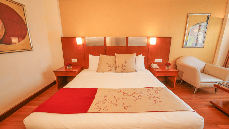 Luxury suite bedroom at The Suryaa New Delhi featuring a large bed with white linens, a beige runner, a red blanket, side tables, and wall-mounted lamps