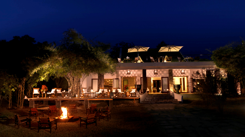 Night view of The Serai Kabini showing a building with outdoor seating on a raised deck, tables with chairs, a fire pit with surrounding chairs in the foreground, trees around the area, and terrace seating on the upper level.