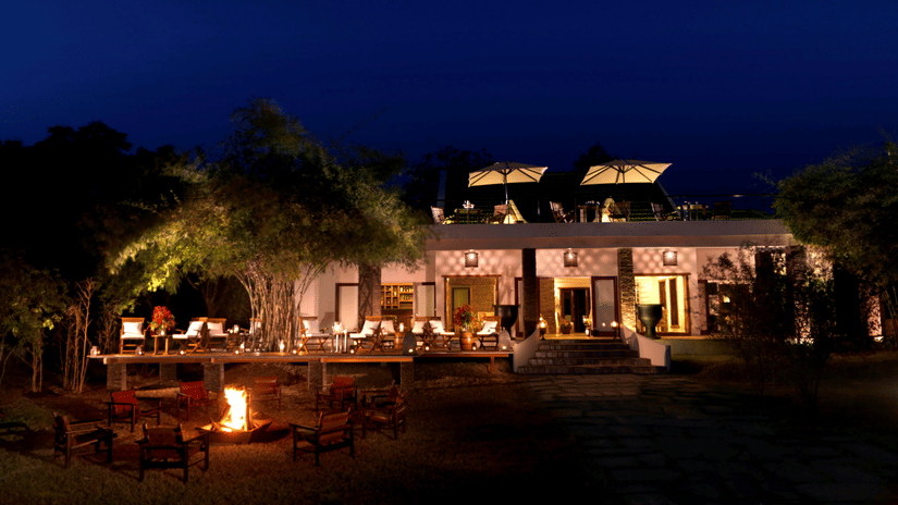Night view of The Serai Kabini showing a building with outdoor seating on a raised deck, tables with chairs, a fire pit with surrounding chairs in the foreground, trees around the area, and terrace seating on the upper level.