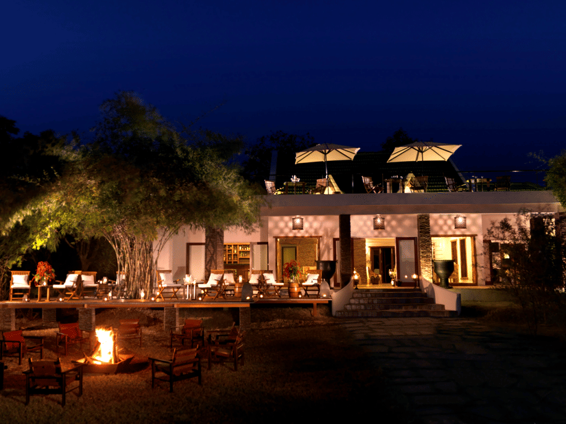 Night view of The Serai Kabini showing a building with outdoor seating on a raised deck, tables with chairs, a fire pit with surrounding chairs in the foreground, trees around the area, and terrace seating on the upper level.