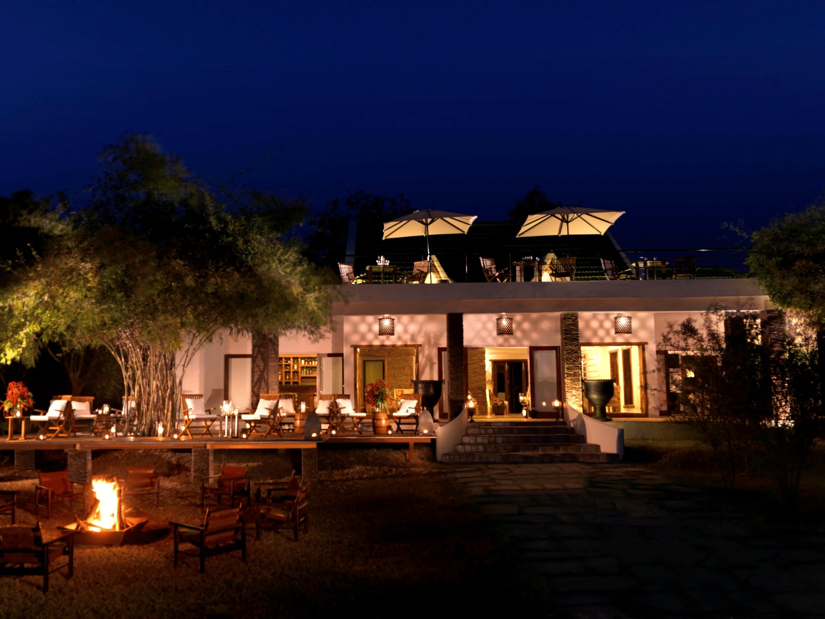 Night view of The Serai Kabini showing a building with outdoor seating on a raised deck, tables with chairs, a fire pit with surrounding chairs in the foreground, trees around the area, and terrace seating on the upper level.