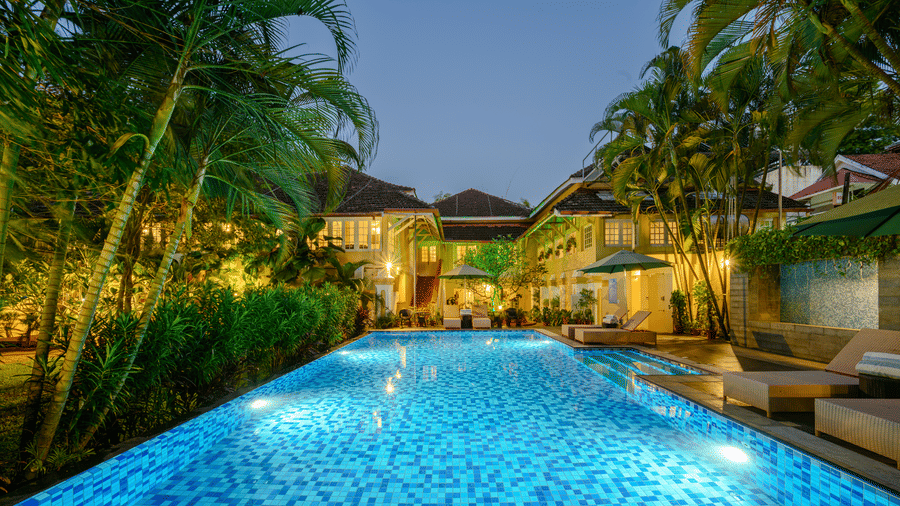 lluminated outdoor swimming pool at night. It is surrounded by lush green palm trees and plants that are lit with warm lighting - a heritage hotel in Kochi The Tower House