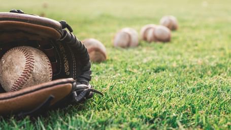 A close-up of a baseball glove with a ball inside, resting on green grass with other baseballs scattered in the background.