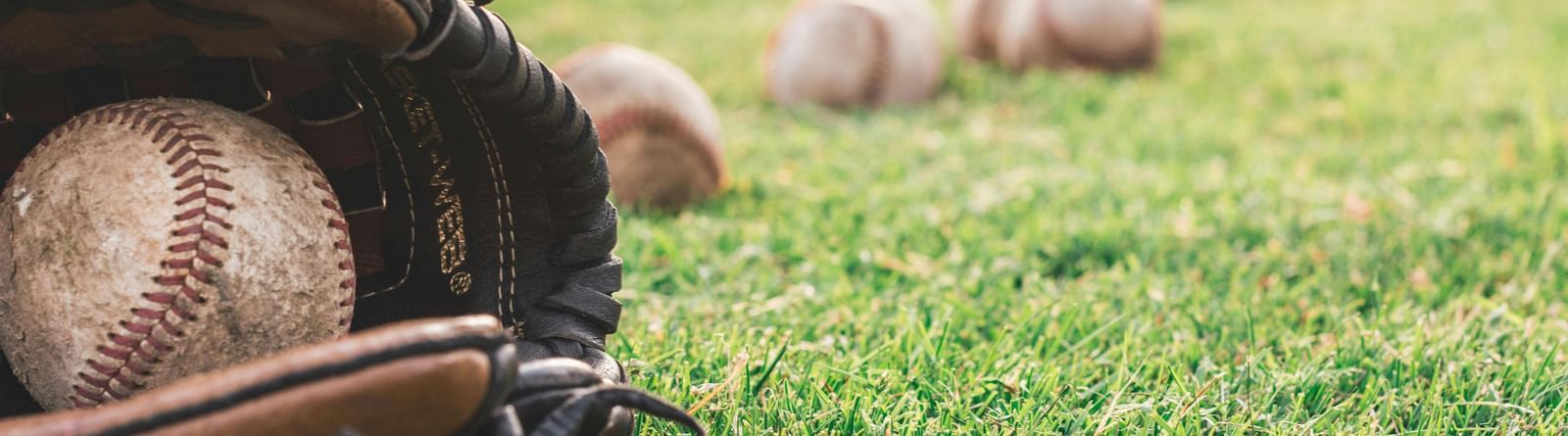 A close-up of a baseball glove with a ball inside, resting on green grass with other baseballs scattered in the background.