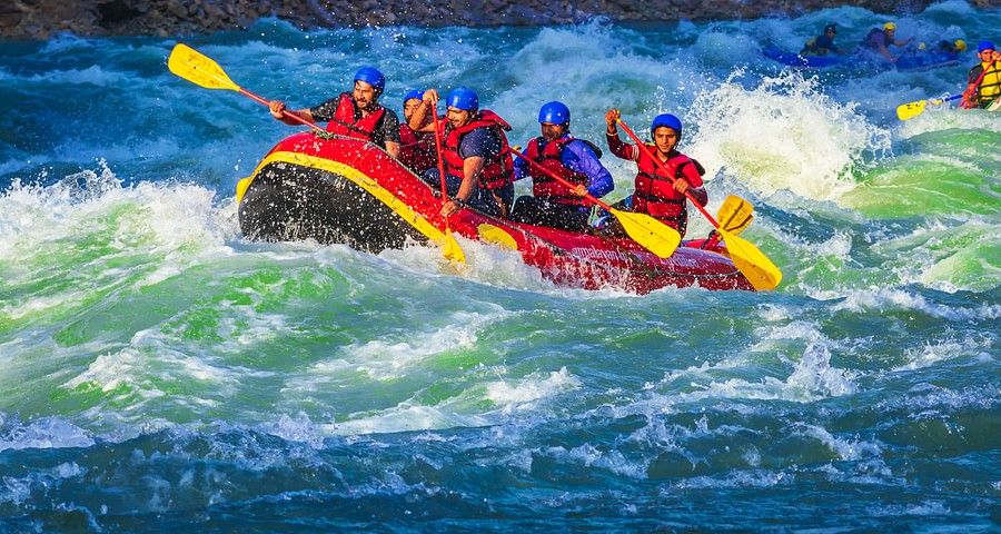 A raft filled with seven people, wearing red life jackets and helmets, is navigating through turbulent, white-capped waters. Everyone appears exhilarated by the action.