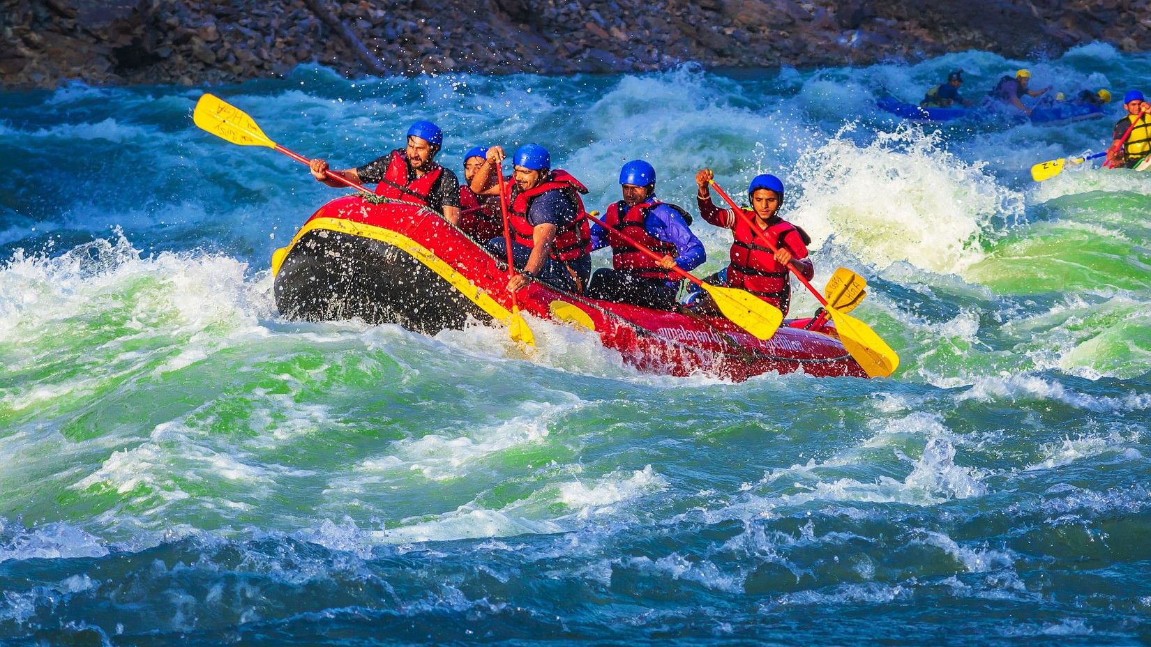 A raft filled with seven people, wearing red life jackets and helmets, is navigating through turbulent, white-capped waters. Everyone appears exhilarated by the action.