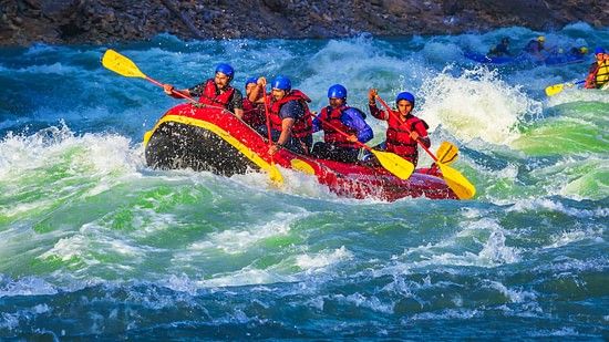 A raft filled with seven people, wearing red life jackets and helmets, is navigating through turbulent, white-capped waters. Everyone appears exhilarated by the action.