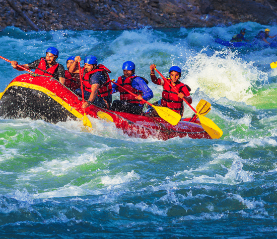 alt-text A raft filled with seven people, wearing red life jackets and helmets, is navigating through turbulent, white-capped waters. Everyone appears exhilarated by the action.