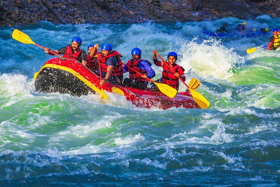 alt-text A raft filled with seven people, wearing red life jackets and helmets, is navigating through turbulent, white-capped waters. Everyone appears exhilarated by the action.