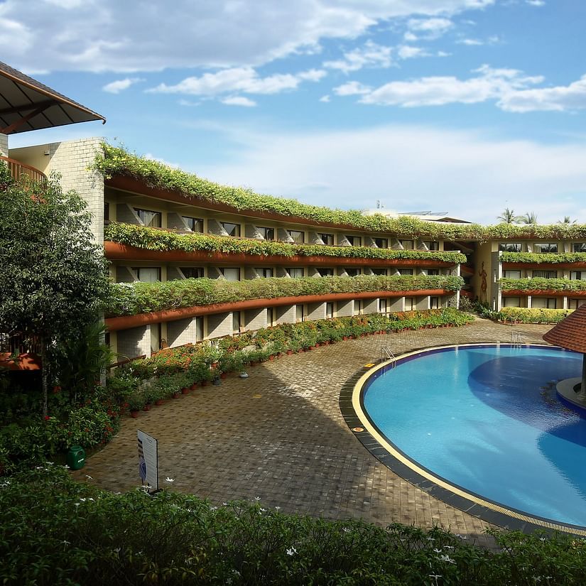 Facade of Uday Suites under a sky with scattered clouds featuring  the hotel building shadowing a large swimming pool