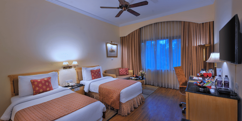A room with twin beds, spreads, pillows, a fan, and windows at The Clarks Varanasi.