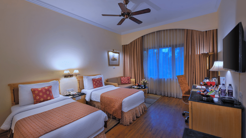 A room with twin beds, spreads, pillows, a fan, and windows at The Clarks Varanasi.