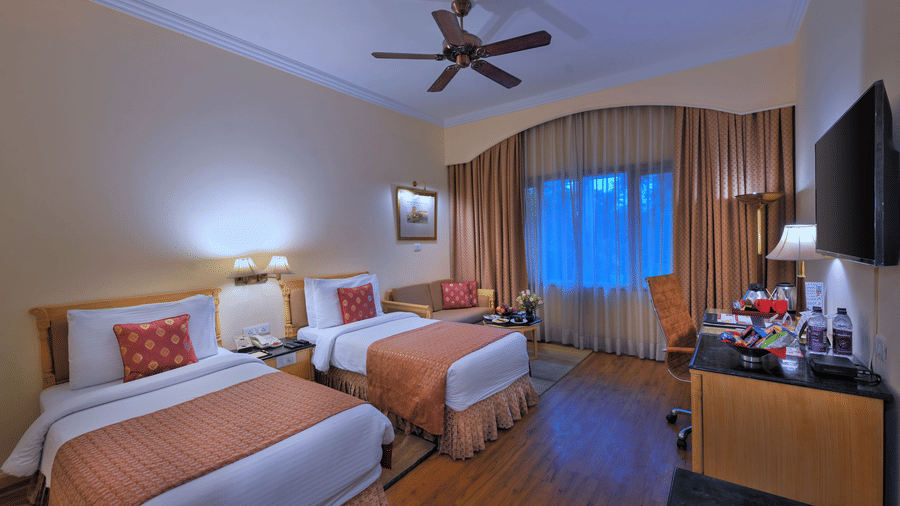 A room with twin beds, spreads, pillows, a fan, and windows at The Clarks Varanasi.