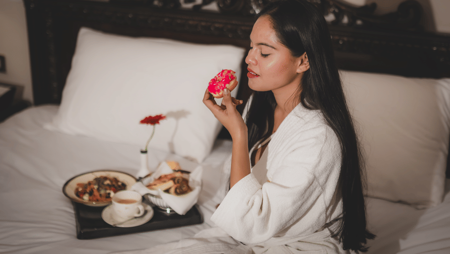 A woman seated on a bed holding a flower and a dining tray with covered dishes placed beside her at The Suryaa, New Delhi.