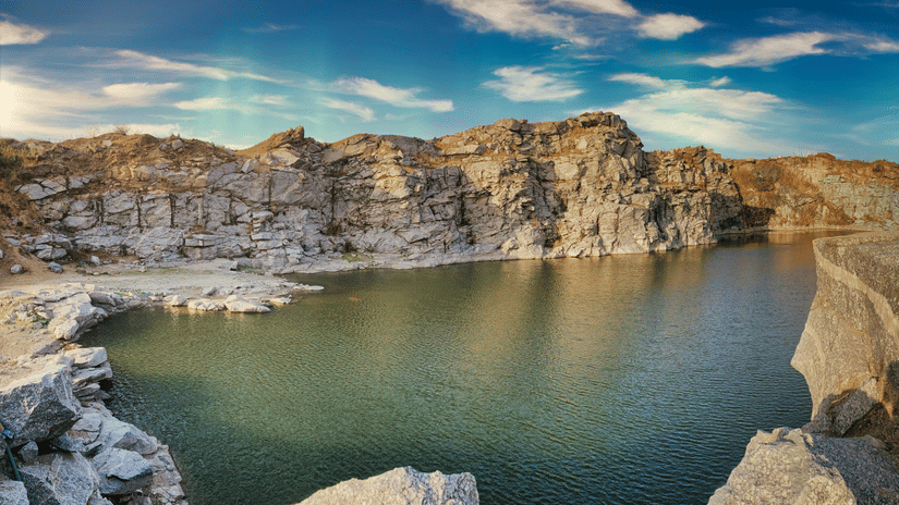 A wide landscape shot of a calm lake at the bottom of a steep, rocky quarry under a bright blue sky with wispy clouds.