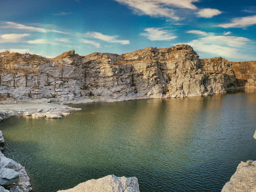A wide landscape shot of a calm lake at the bottom of a steep, rocky quarry under a bright blue sky with wispy clouds.