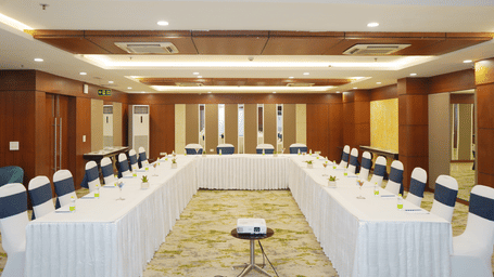 A meeting room configured with a U-shaped table layout, white tablecloths, and an overhead wooden ceiling feature.
