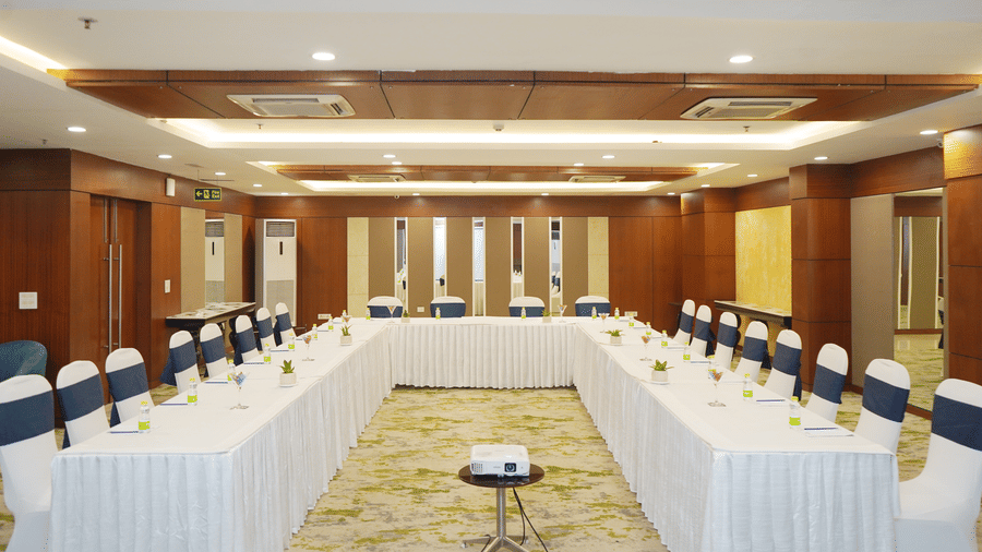 A meeting room configured with a U-shaped table layout, white tablecloths, and an overhead wooden ceiling feature.