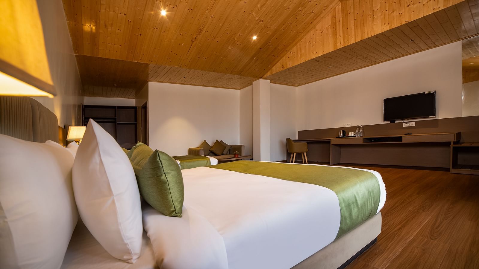 Modern hotel room featuring a cosy bed with green accents, wooden flooring, a sleek work desk with a television, and a stylish wooden ceiling with warm lighting - Yashshree Pelling Chekhim Retreat.