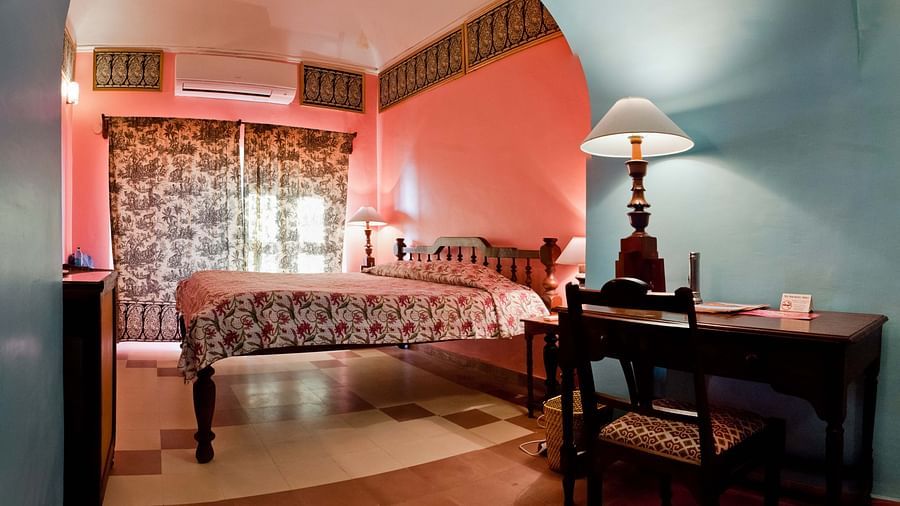 Bedroom with a bed featuring a floral cover, bedside tables with lamps, a desk and chair, patterned curtains, and AC at Tijara Fort-Palace, Alwar.